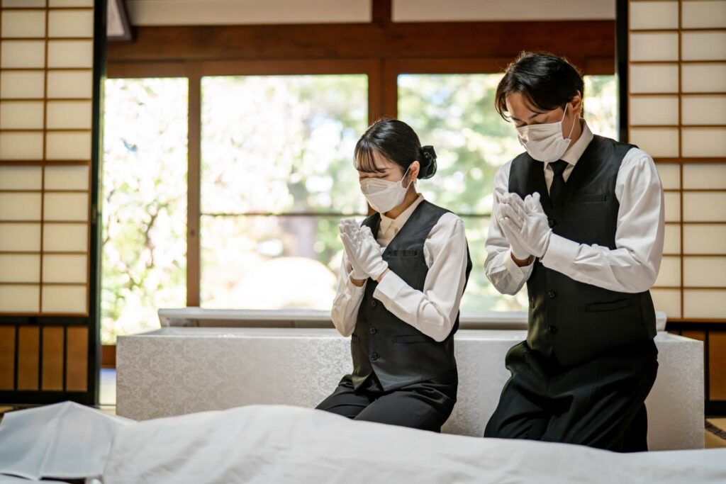 マスクと白手袋をつけて遺体の前で手を合わせる葬儀社スタッフの男女